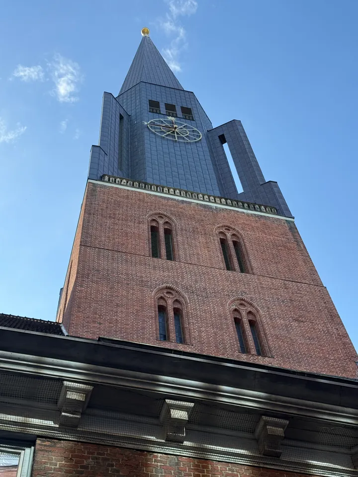 Kirchturm von unten gesehen: Backsteinschaft mit Rundbogenfenstern, dunkler Turmhelm mit Uhr und goldener Spitze