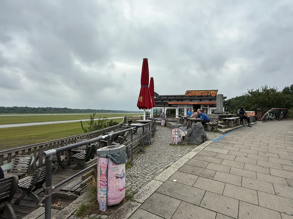 Café „Coffee to Fly“ mit Außensitzplätzen, Sonnenschirmen und Pflasterweg neben der Flughafenwiese