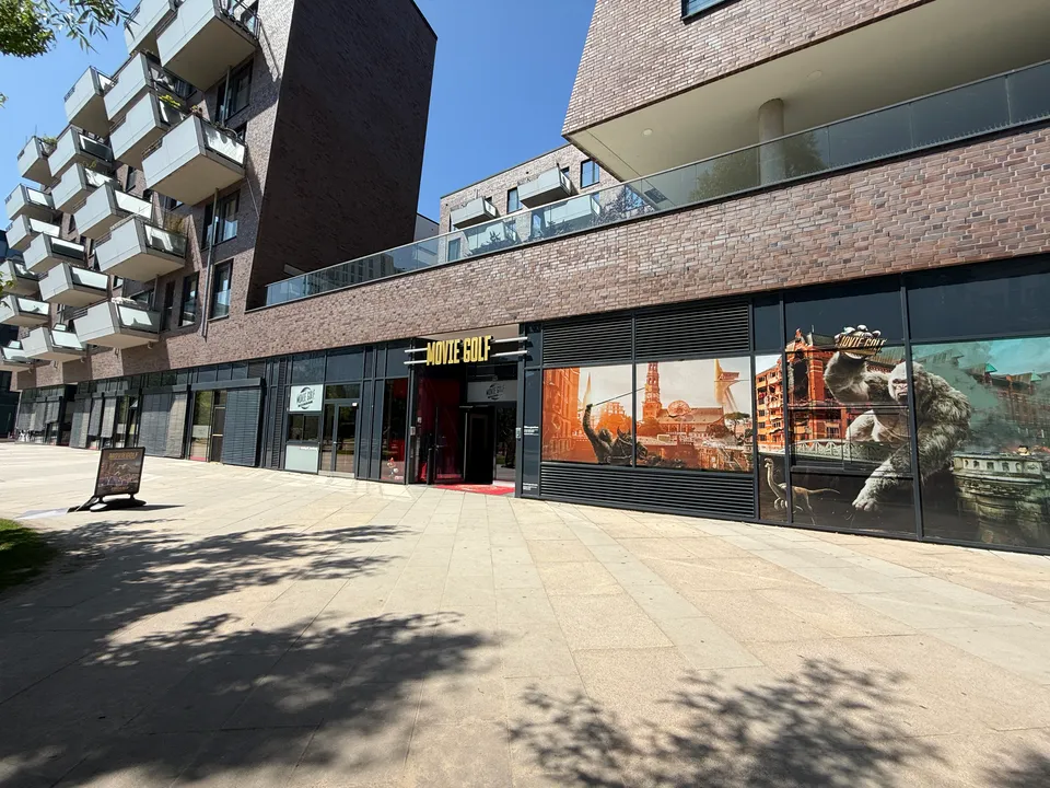 Klinkerbau mit Balkonen, im Erdgeschoss Eingang von Movie Golf und bunt beklebte Schaufenster am Gehweg