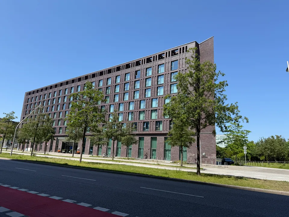 Langgestrecktes achtgeschossiges Klinkergebäude mit spitzer Ecke hinter Straßenbäumen, im Vordergrund Straße und roter Radweg