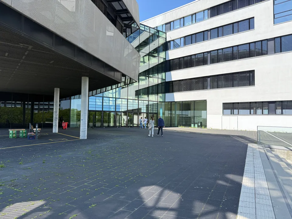 Gepflasterter Platz vor verglastem Eingang mit diagonaler Treppenhausfassade, links auskragender Gebäudeteil auf Betonstützen