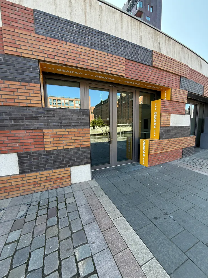 Glastür des Pavillons OSAKA9 mit gelbem Rahmen in einer Fassade aus verschiedenfarbigen Klinkerfeldern