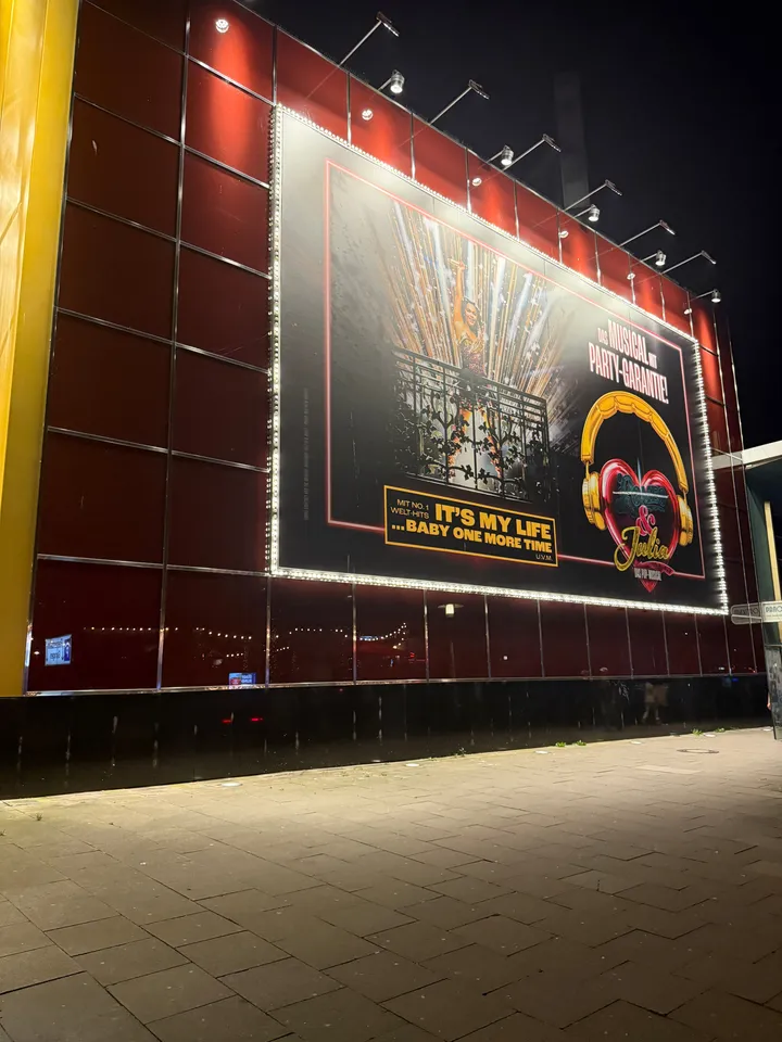 Rote Theaterfassade bei Nacht mit großem beleuchtetem Musicalplakat für „& Julia“ und leerem Gehweg davor