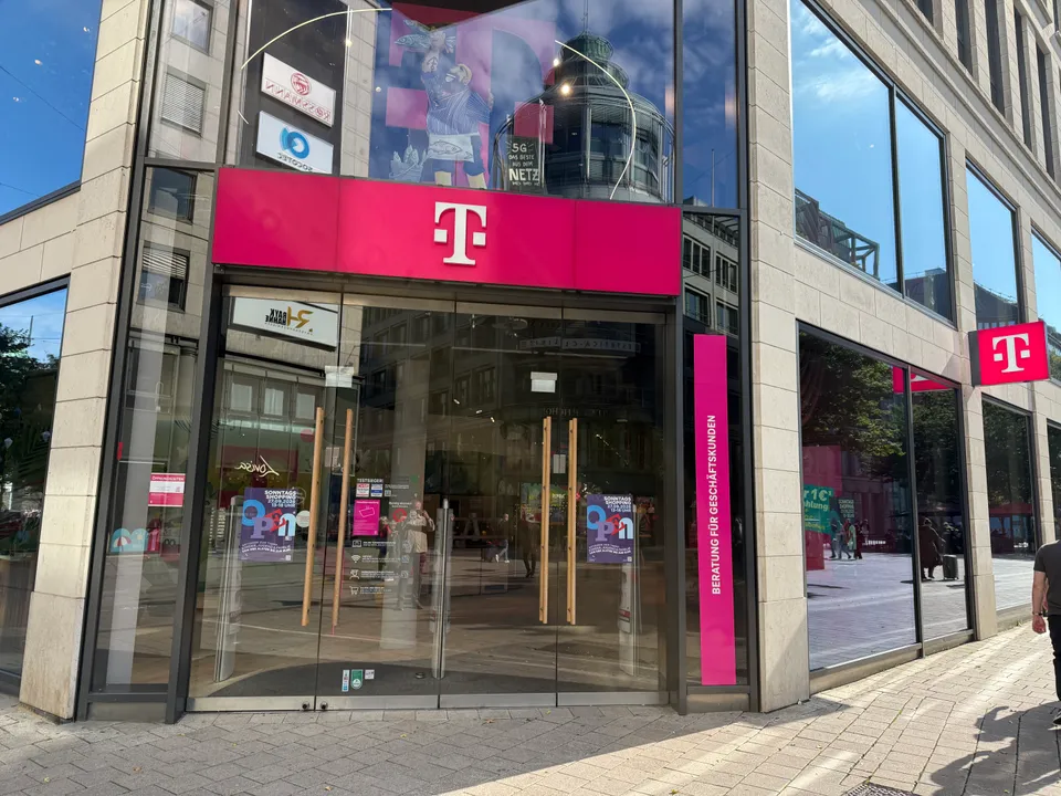 Eingang eines Telekom Shops mit magentafarbenem T-Logo und Glastüren in einer sonnigen Fußgängerzone
