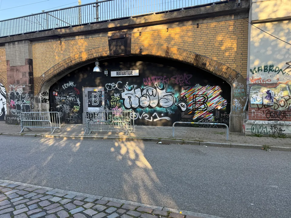 Mit Graffiti bedeckte schwarze Front in einem Klinker-Viaduktbogen mit Schild „Beatboutique“, davor Absperrgitter auf dem Gehweg