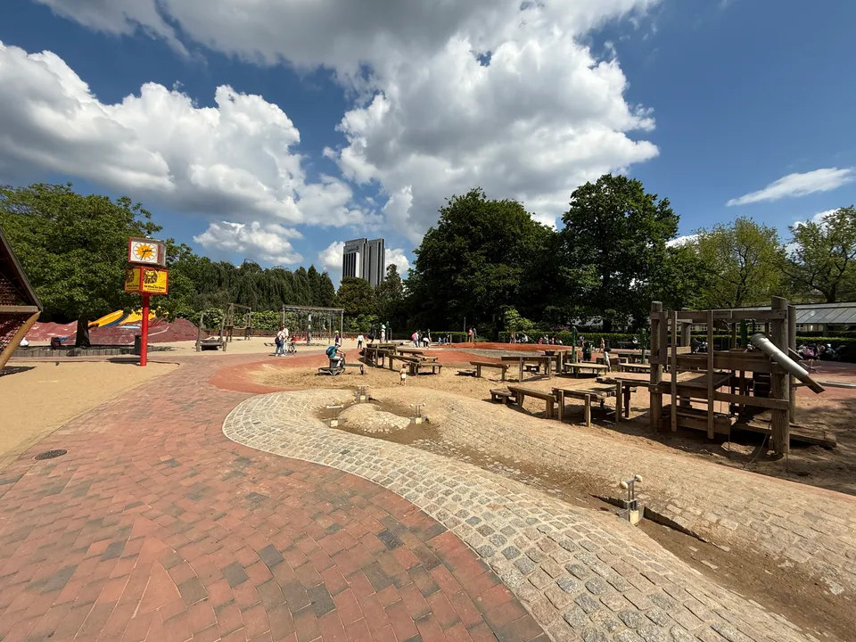 Gepflasterte Wasserrinne und Matschspielbereich mit Holztischen, dahinter Bäume und ein Hochhaus