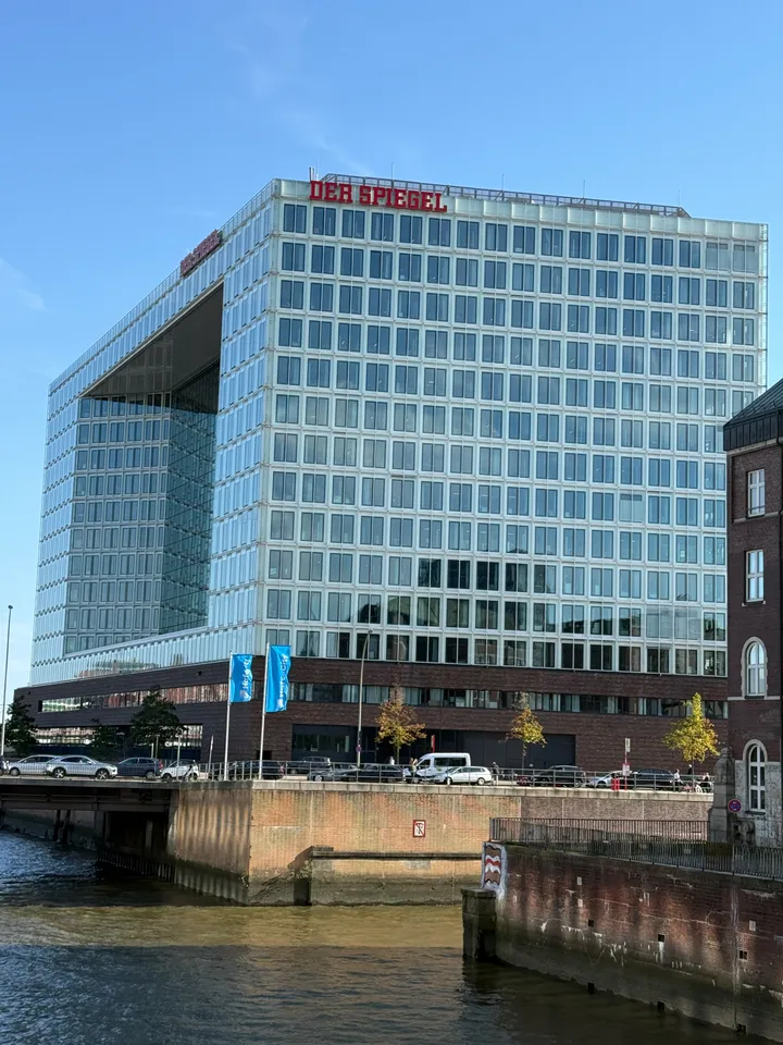 SPIEGEL-Gebäude mit roter Dachschrift hinter einem Kanal, Brücke und Kaimauer, rechts ein Backsteinbau