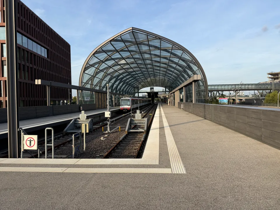 Gewölbte Glashalle eines Hochbahnhofs mit einfahrbereitem U-Bahn-Zug, Prellböcken und leerem Bahnsteig unter blauem Himmel