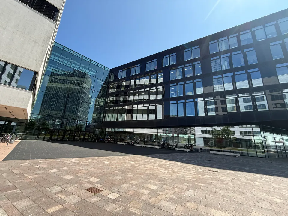 Gepflasterter Vorplatz vor Hochschulgebäude mit verglastem Eingangsbereich und dunklem, auskragendem Fassadenriegel bei blauem Himmel