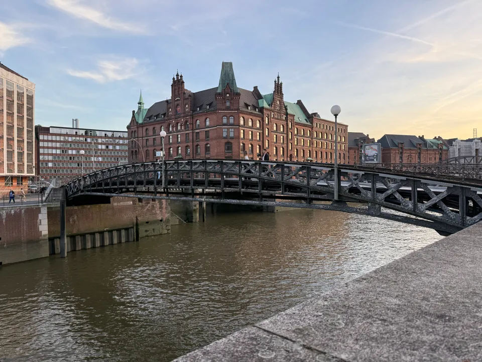Genietete Stahlbogenbrücke über einen Kanal vor einem neugotischen Backsteingebäude mit grünen Kupferdächern im Abendlicht