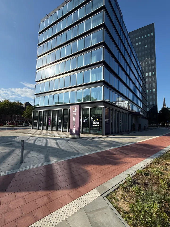 Mehrgeschossiges Hotel mit Glasfassade und Premier-Inn-Stele, dahinter dunkles Hochhaus, davor roter Radweg unter blauem Himmel