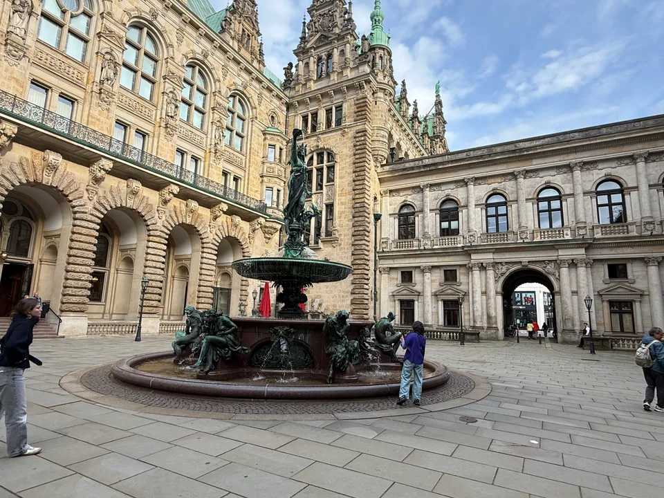 Bronzebrunnen mit Frauenfigur und sitzenden Figuren im gepflasterten Innenhof vor verzierter Sandsteinfassade mit Turm