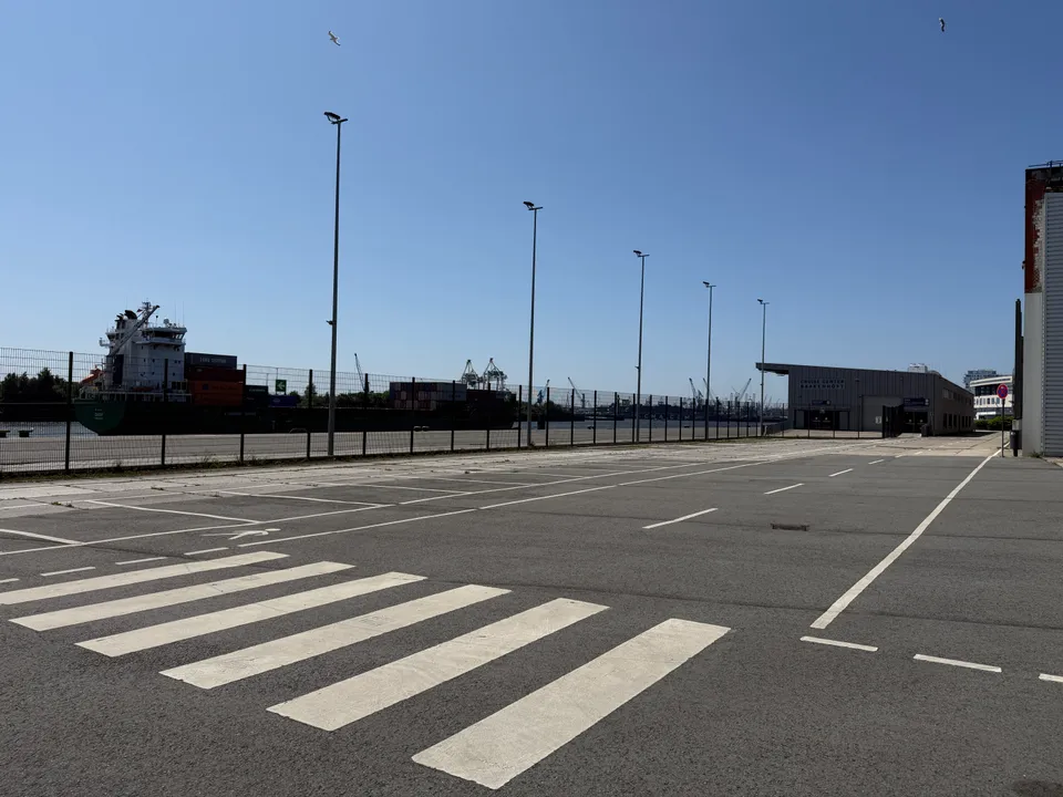Leere Parkfläche mit Zebrastreifen und Lichtmasten, links Containerschiff hinter Zaun, hinten Terminalhalle