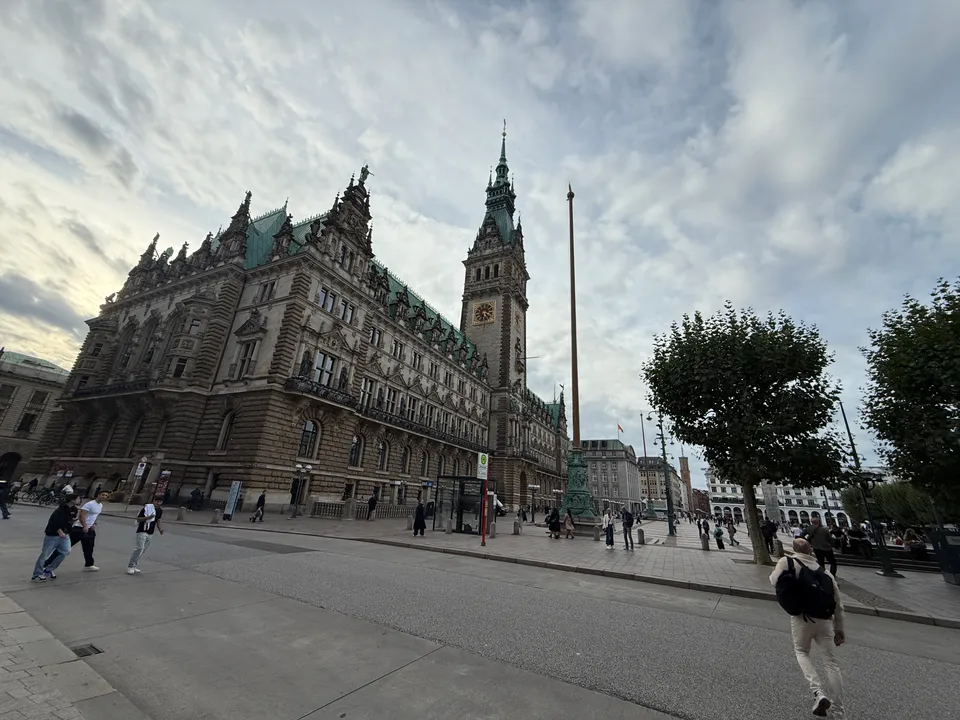 Hamburger Rathaus mit Uhrturm am Rathausmarkt, davor Fahnenmast, Bäume und Passanten auf der Straße