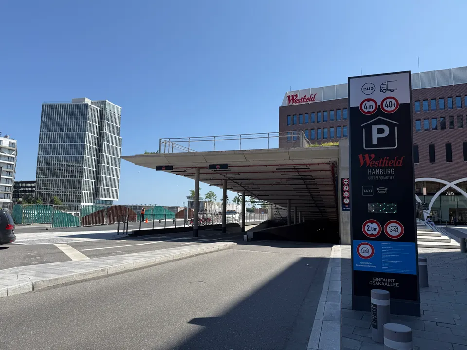 Tiefgarageneinfahrt Osakaallee des Westfield Hamburg-Überseequartier mit Parkhaus-Stele, Klinkerbau und Glashochhaus bei blauem Himmel