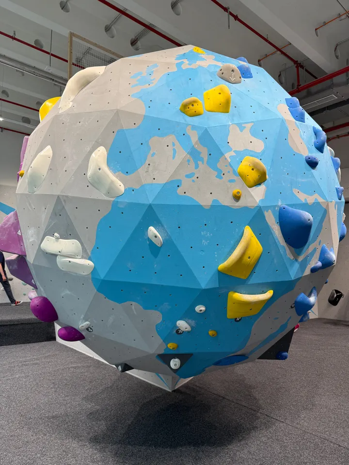 Kugelförmige Kletterstruktur als Globus bemalt mit bunten Griffen über grauer Matte in einer Boulderhalle