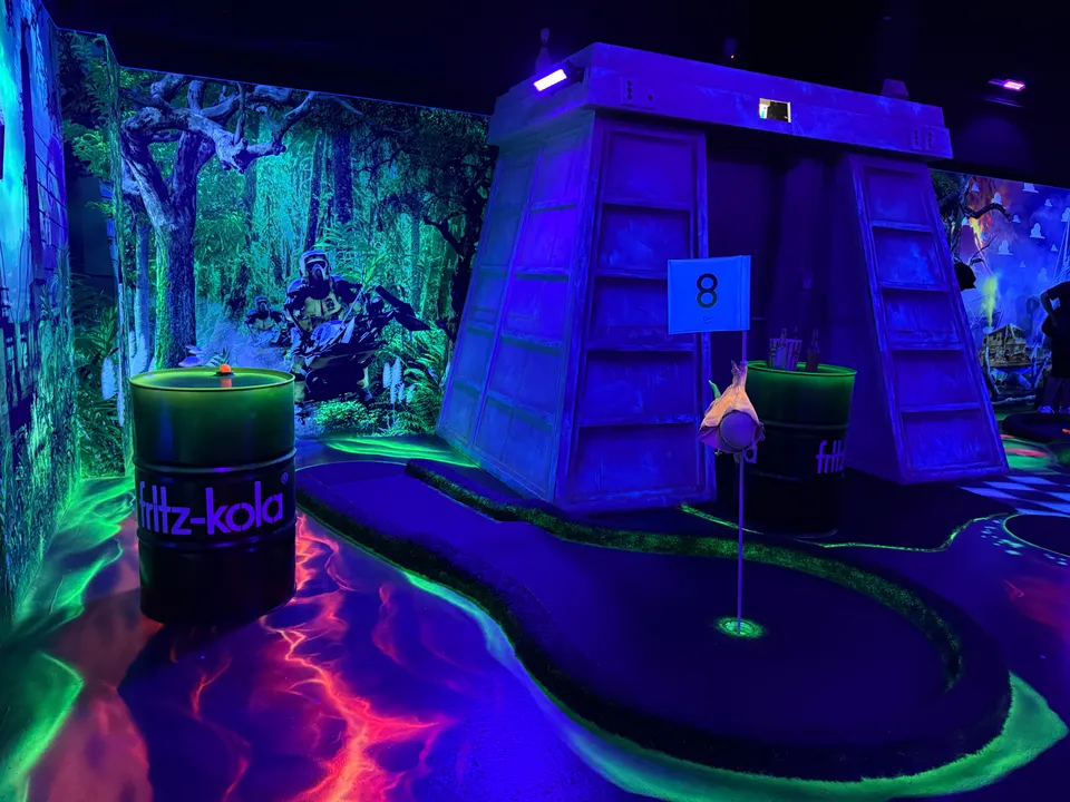 Minigolfbahn 8 im Schwarzlicht vor Waldkulisse mit bunkerartigem Aufbau und zwei leuchtenden Fässern