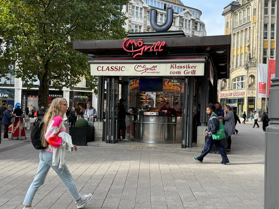 Imbisspavillon MöGrill mit U-Bahn-Symbol auf dem Dach in einer Fußgängerzone, davor gehen Passanten vorbei