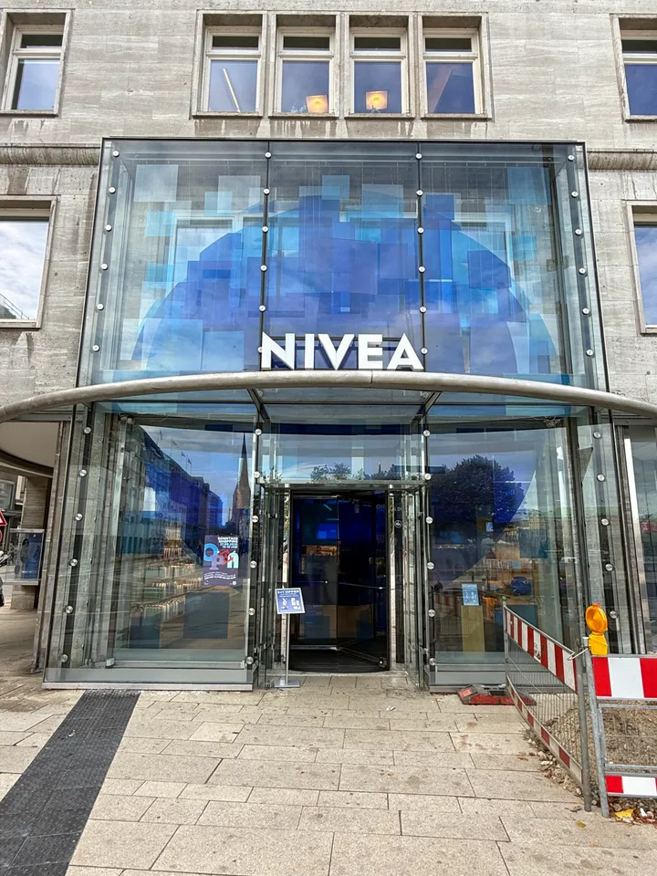 Verglaster Eingang mit weißem NIVEA-Schriftzug auf blauem Glasmosaik, Drehtür und rot-weißen Absperrgittern daneben