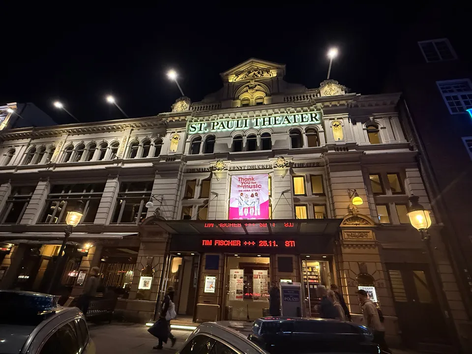 Beleuchtete Fassade des St. Pauli Theaters bei Nacht mit ABBA-Banner, Laufschrift und parkenden Autos davor