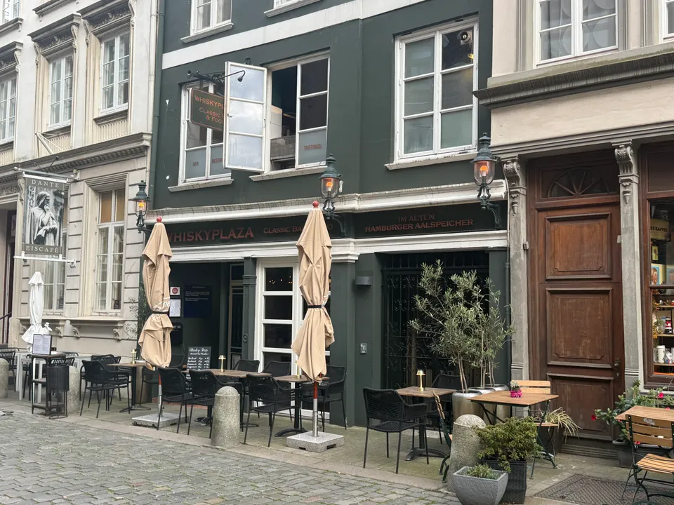 Dunkelgrüne Altbaufassade der Whiskyplaza mit Außentischen, zugeklappten Sonnenschirmen und Laternen an gepflasterter Straße