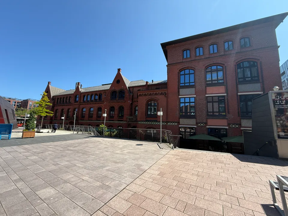 Historisches Backsteingebäude mit neugotischen Giebeln und höherem Eckbau an einem weiten, gepflasterten Platz unter blauem Himmel