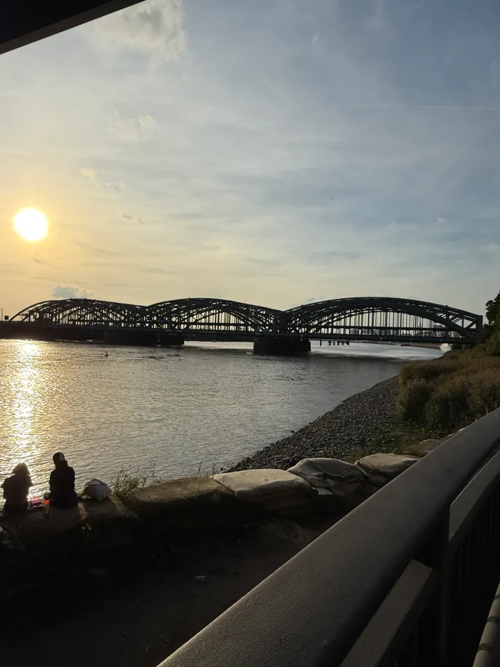 Dreibogige Stahlbrücke über der Elbe bei tiefstehender Sonne, davor Kiesufer mit Sandsäcken und Geländer