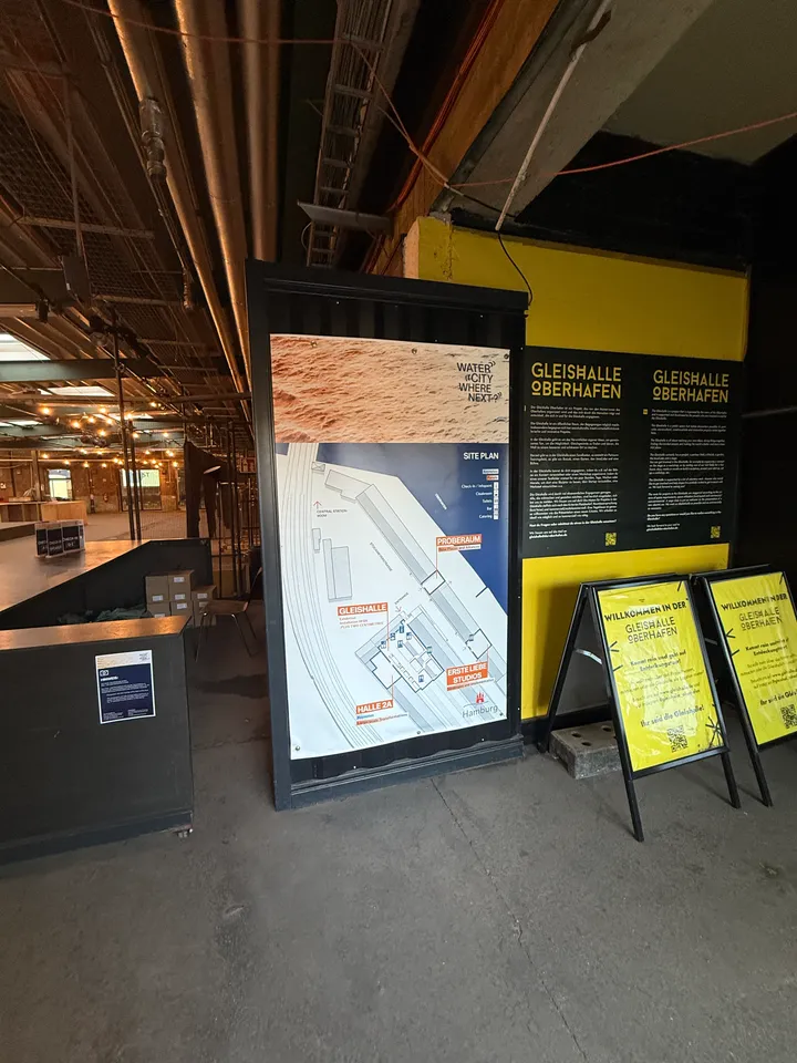 Lageplan, gelbe Infotafel und zwei Aufsteller mit Begrüßungstext im Eingangsbereich der Halle