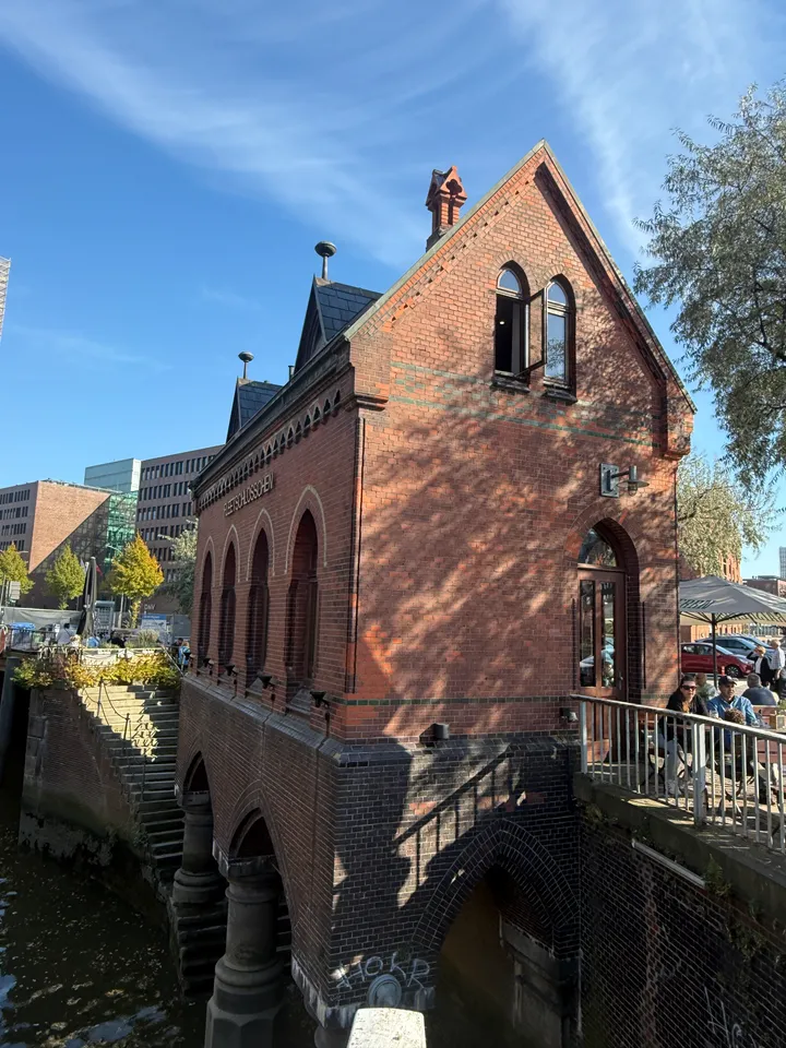 Schmales neugotisches Backsteinhaus mit Spitzgiebel auf Bögen über einem Fleet, rechts Gäste an einer Terrasse
