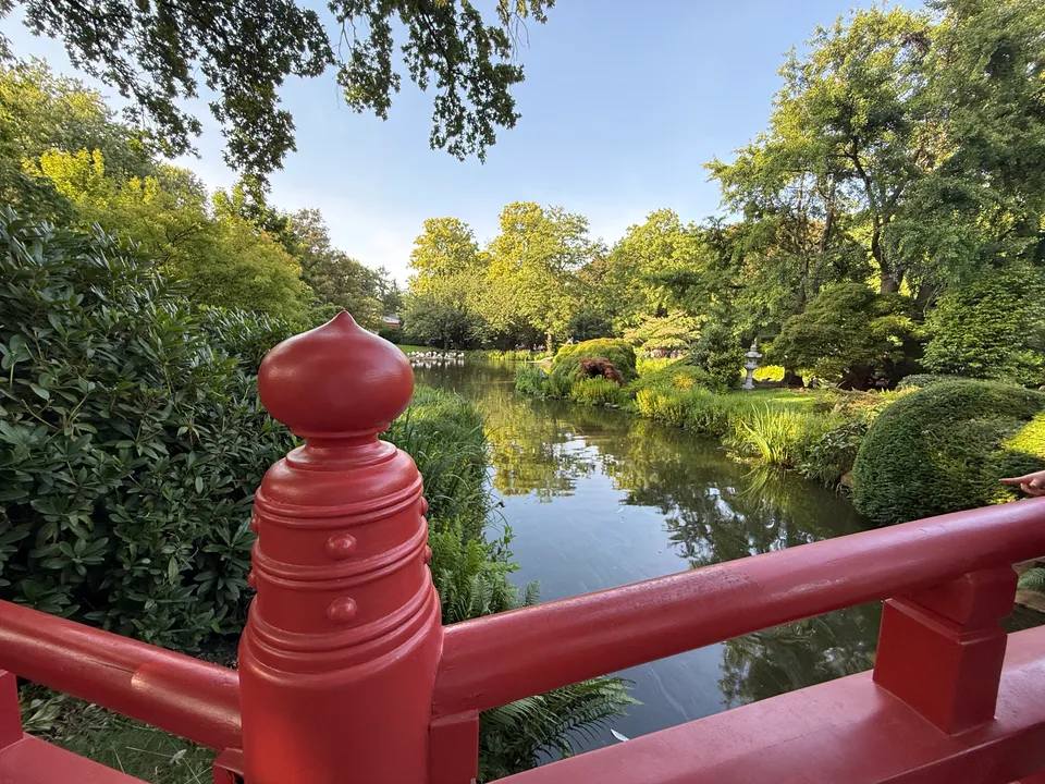 Rote Brücke im japanischen Stil über einem Teich, gesäumt von Büschen, Bäumen und einer Steinlaterne