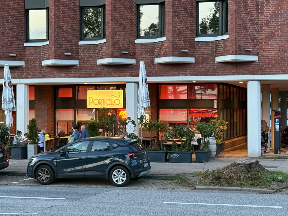 Backsteingebäude mit Restaurant „Ristorante Porticello“ im Erdgeschoss, Außenterrasse mit Pflanzkübeln, davor ein geparktes dunkles Auto