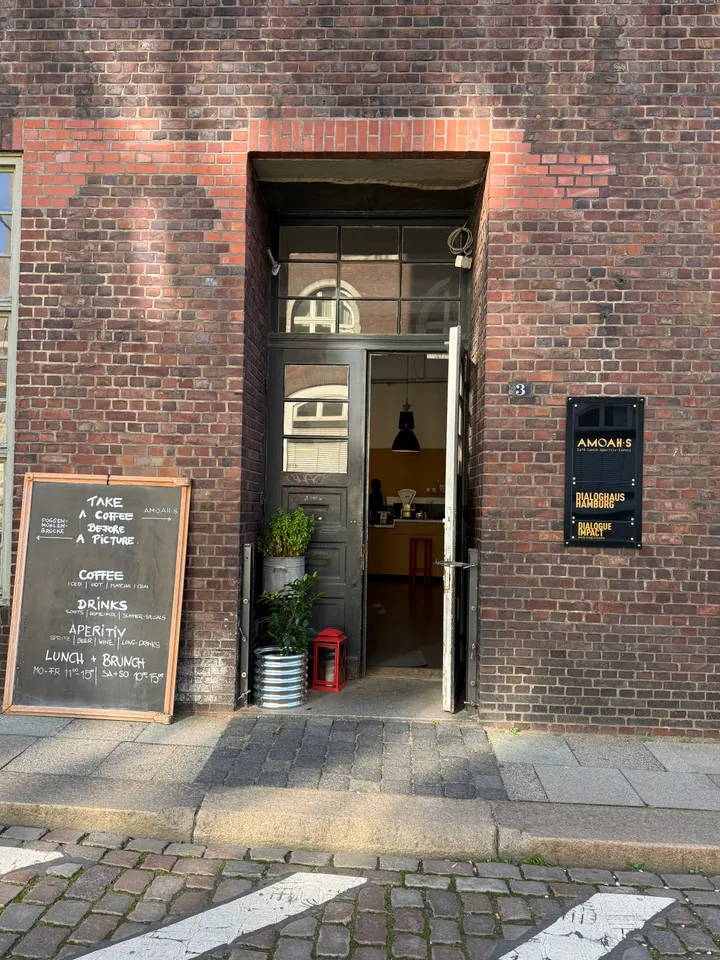Offene Eingangstür des Cafés AMOAHS in Backsteinfassade mit Kreidetafel, Pflanzkübeln und schwarzem Hinweisschild