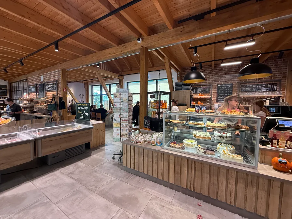 Hofladen mit Holzdachstuhl, Tortenvitrine an der Cafétheke, Kühltheken und Sitzbereich im Hintergrund