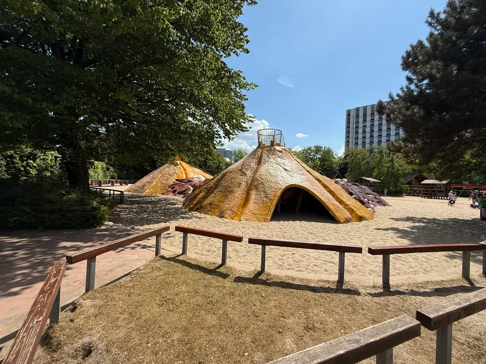 Gelbe kegelförmige Kletterberge mit Durchgang auf großer Sandfläche, dahinter Bäume und ein Hochhaus