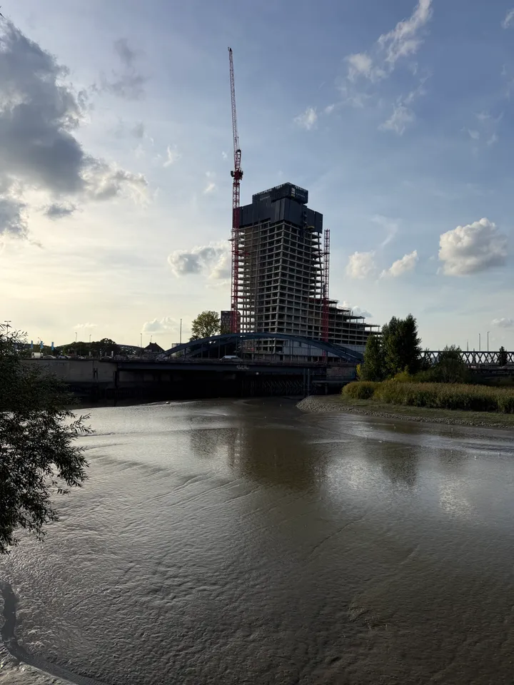 Wasserfläche mit Spiegelungen vor einer blauen Bogenbrücke und einem Hochhausrohbau mit rotem Kran