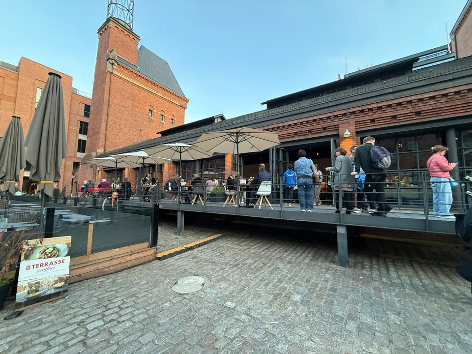 Backsteingebäude mit Turm und erhöhter Restaurantterrasse mit Sonnenschirmen und Gästen über einem Platz mit Kopfsteinpflaster