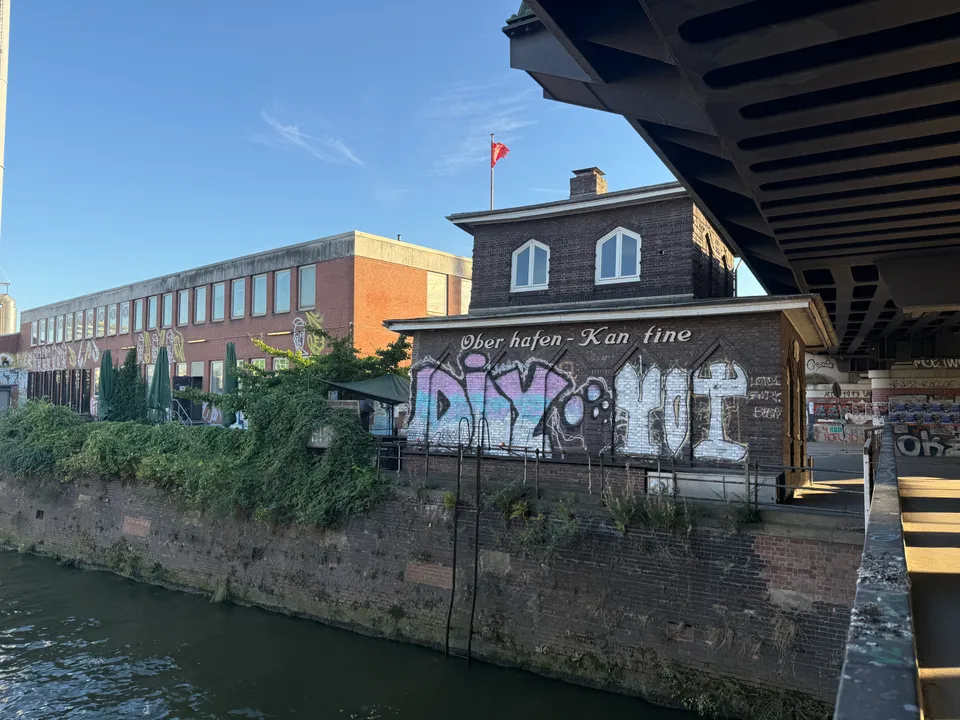 Graffiti-bemalte Oberhafen-Kantine an einer Kaimauer neben einer Stahlbrücke, links ein Backstein-Bürogebäude