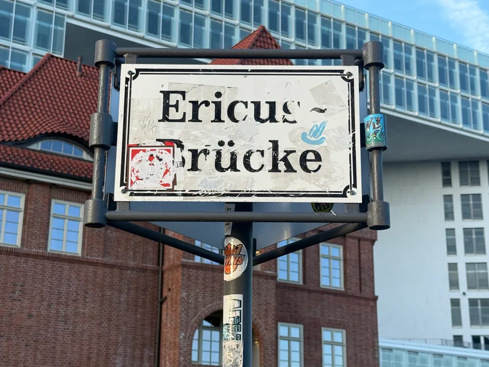 Straßenschild „Ericus-Brücke“ mit Aufklebern vor einem Backsteingebäude mit rotem Dach und einem modernen Glasbau