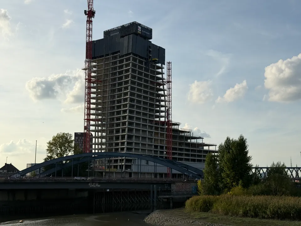 Hochhausrohbau mit Aufschrift Elbtower hinter blauer Bogenbrücke, davor Kiesufer, Schilf und Bäume