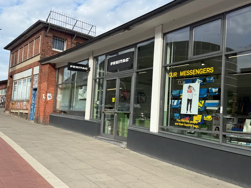 Ladenfront des FREITAG Stores mit Schaufenster voller bunter Umhängetaschen, daneben ein altes Backsteingebäude am Gehweg