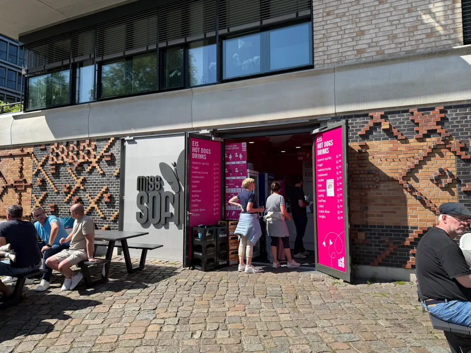Geöffneter Kiosk Miss Sofie mit pinken Preistafeln, Klinkerfassade und Gästen an Picknicktischen auf Kopfsteinpflaster