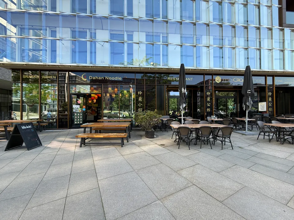 Glasfassade mit Restaurant Dahan Noodle im Erdgeschoss, davor leere Außenterrasse mit Holzbänken und Bistrotischen