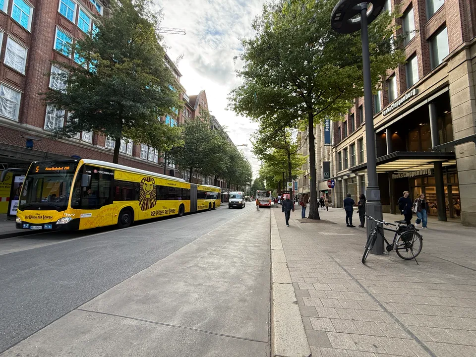 Gelber Gelenkbus auf baumgesäumter Einkaufsstraße, rechts Kaufhaus Peek & Cloppenburg, Fahrrad am Laternenmast und Fußgänger
