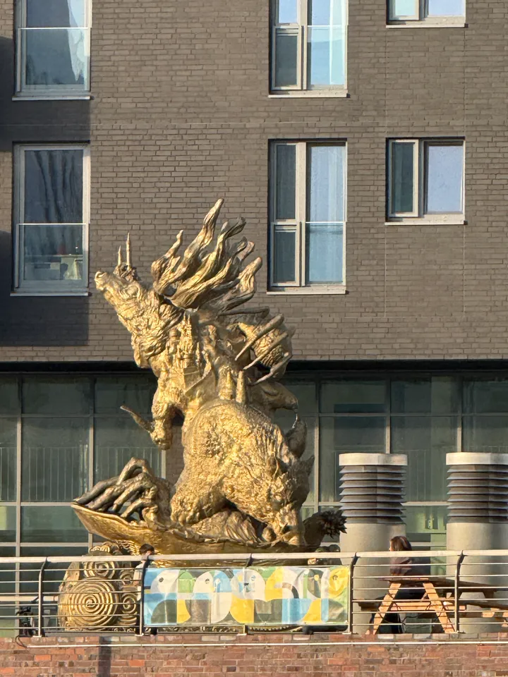 Goldene Skulptur aus verschlungenen Wildtierfiguren auf einer Terrasse hinter einem Geländer mit buntem Banner