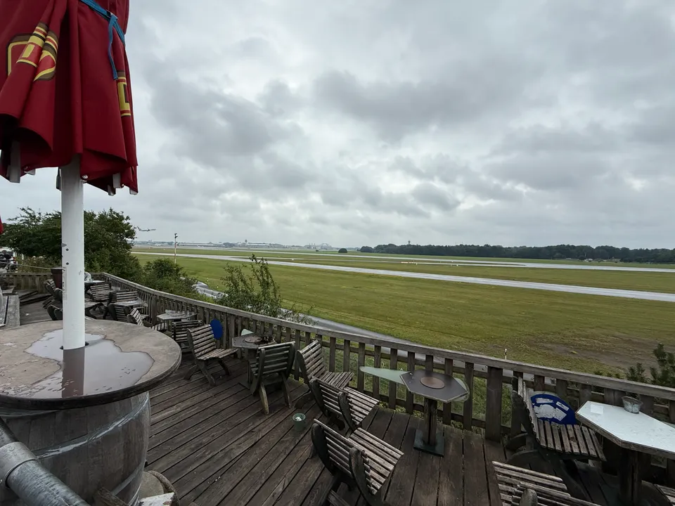Leere Holzterrasse mit Stühlen und Sonnenschirm, Blick auf Start- und Landebahn unter bewölktem Himmel