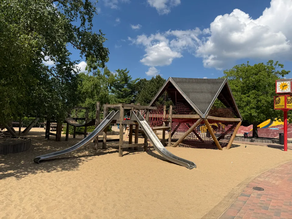 Holzspielturm mit zwei Metallrutschen und dachförmigem Netzklettergerüst auf einer Sandfläche