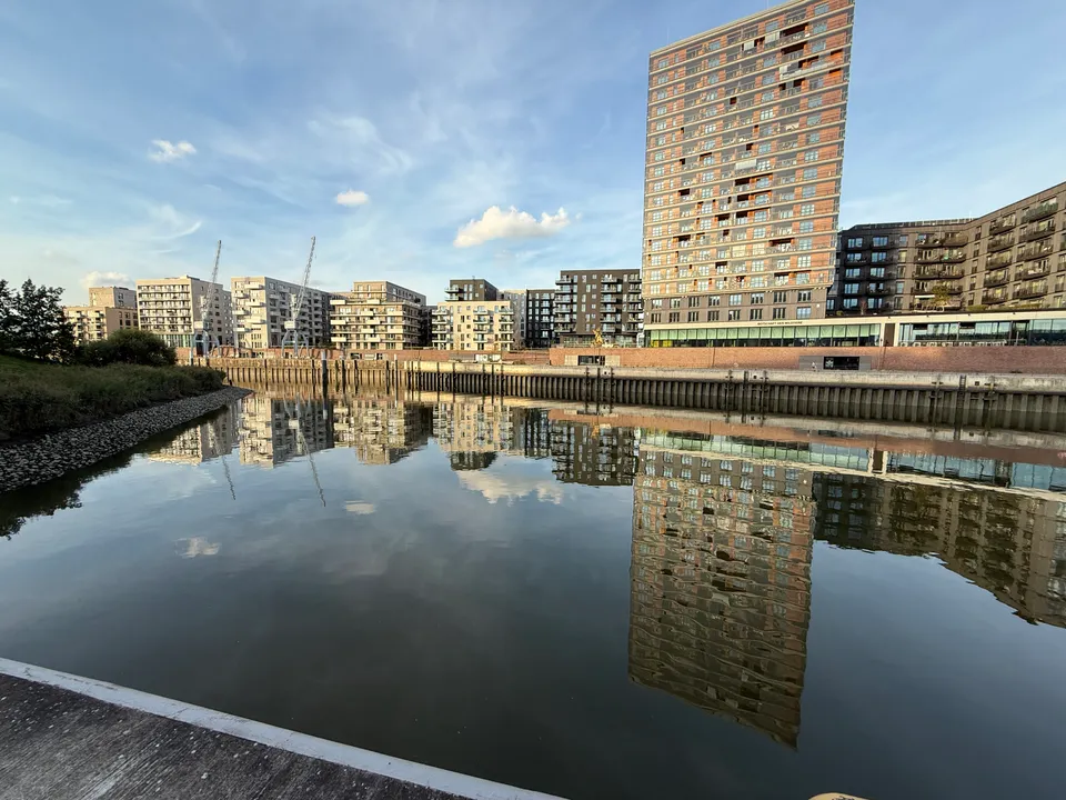 Wohnhochhaus mit rötlicher Fassade und Neubauten am Hafenbecken, die sich im ruhigen Wasser spiegeln