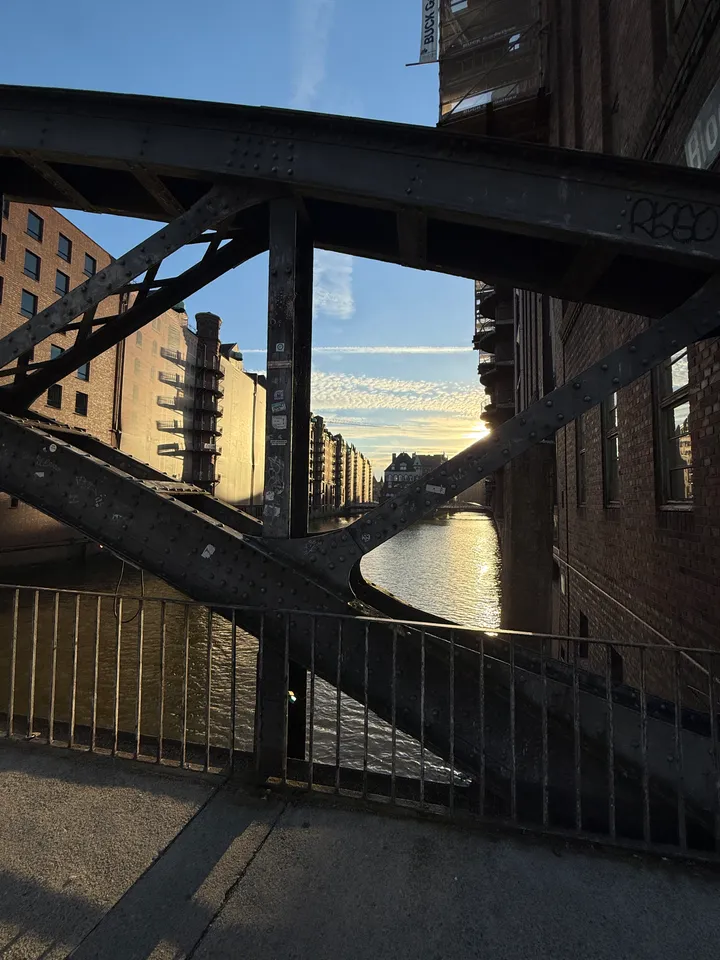 Genietete Stahlstreben einer Brücke, dahinter ein sonnenbeschienenes Fleet mit Speicherzeilen und dem Wasserschloss in der Ferne