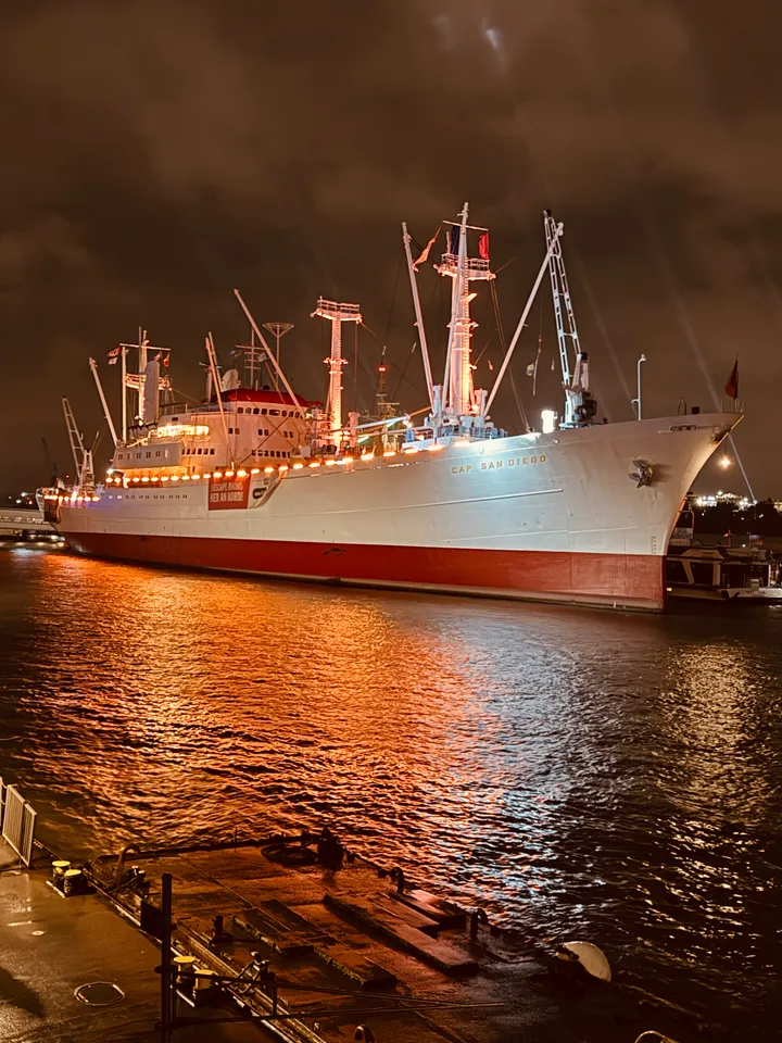 Angestrahltes weißes Museumsschiff Cap San Diego mit rotem Rumpf nachts am Kai, Lichter spiegeln sich im Wasser