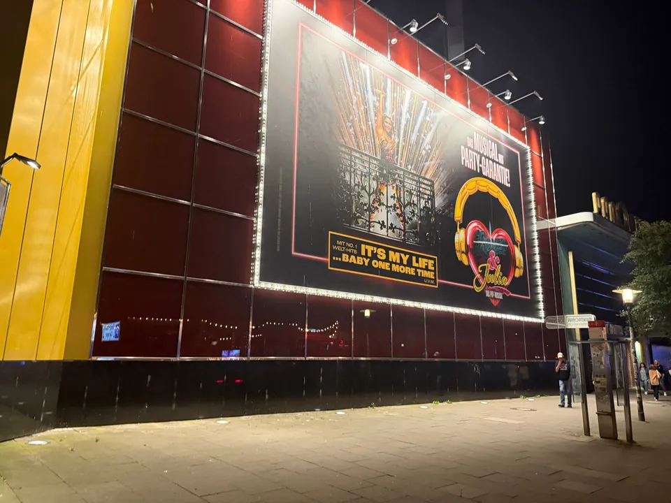 Rote Theaterfassade mit beleuchtetem Musicalplakat bei Nacht, rechts Straßenlaterne und Eingang des Nachbargebäudes